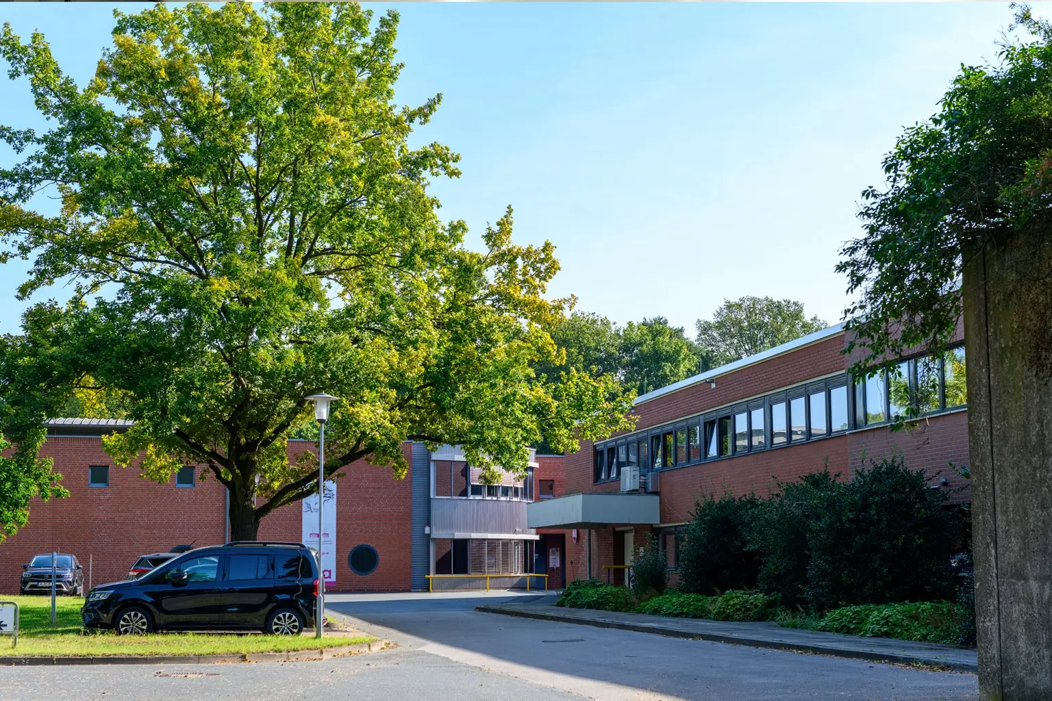 Außenansicht eines Backsteingebäudes mit grünen Bäumen und einer Zufahrt.