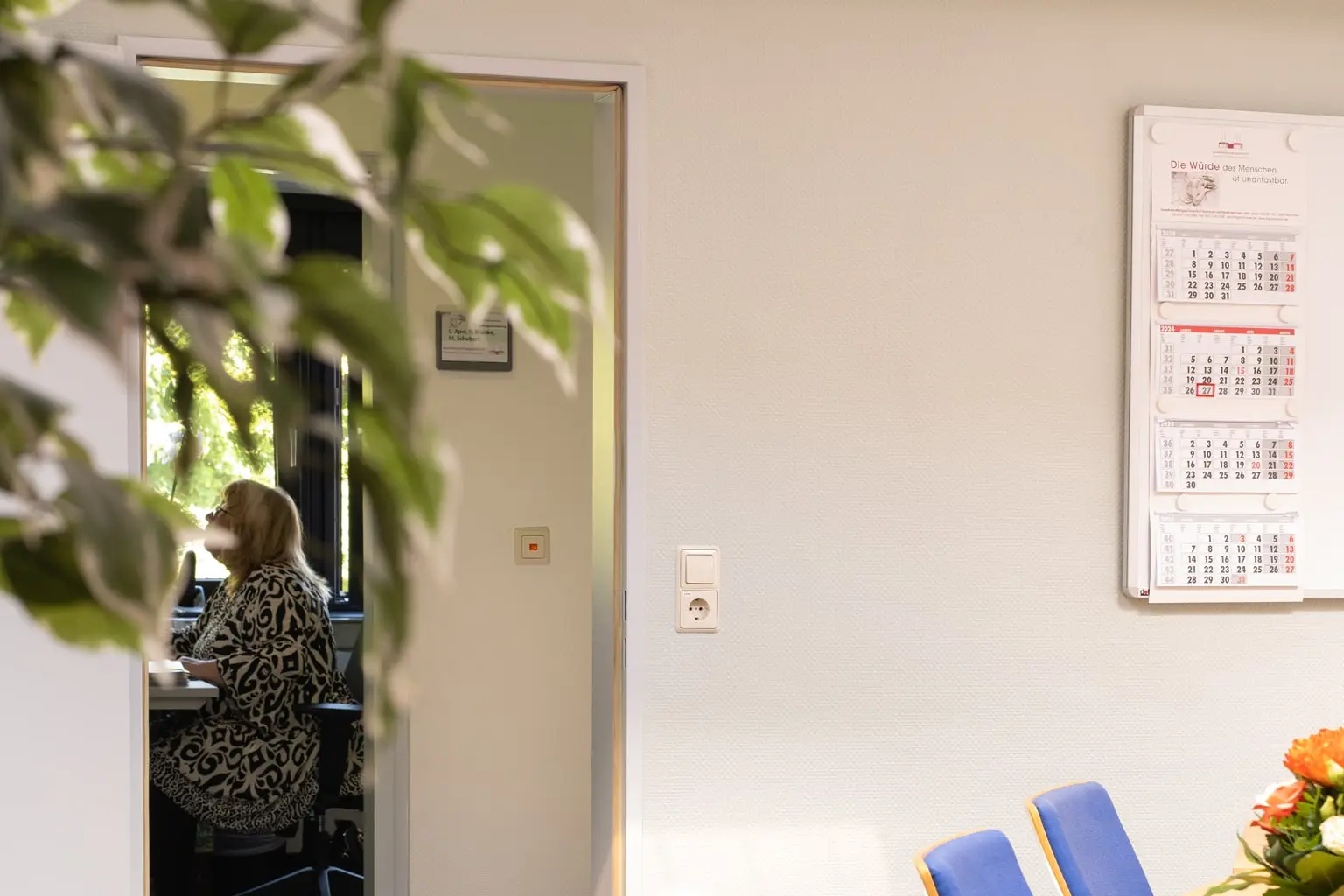 Bürobereich mit einer am Schreibtisch sitzenden Frau, einer Pflanze und Kalendern an der Wand.