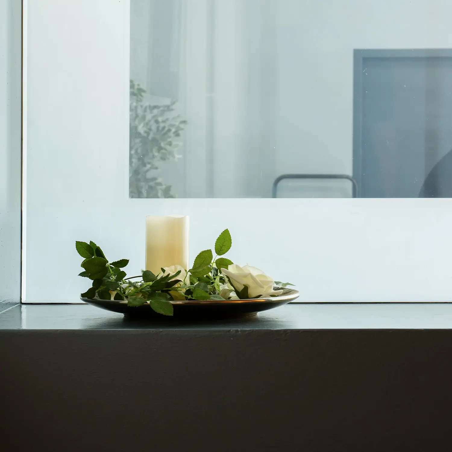 Weiße Kerze, Rosen und grünes Laub auf einem Dekoteller vor hellem Hintergrund.