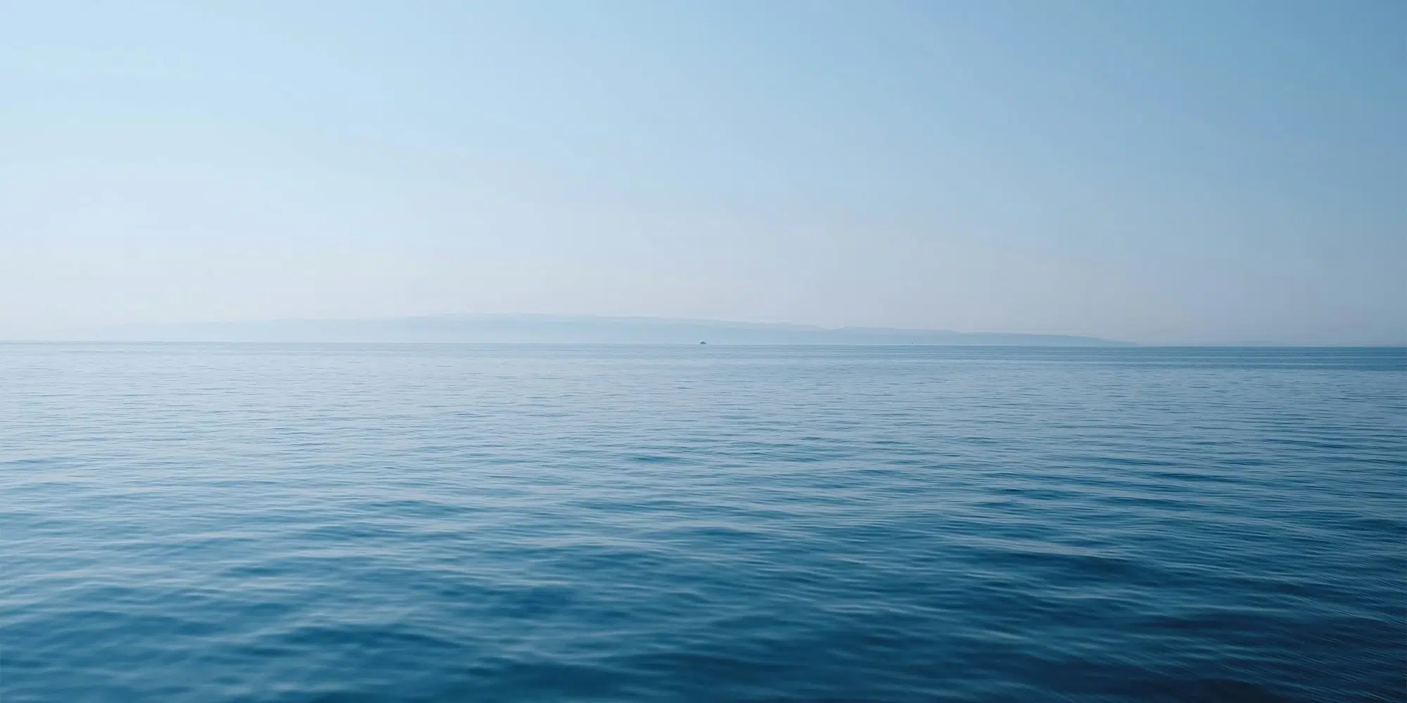 Ruhiges blaues Meer unter hellem Himmel, am Horizont eine ferne Landmasse.