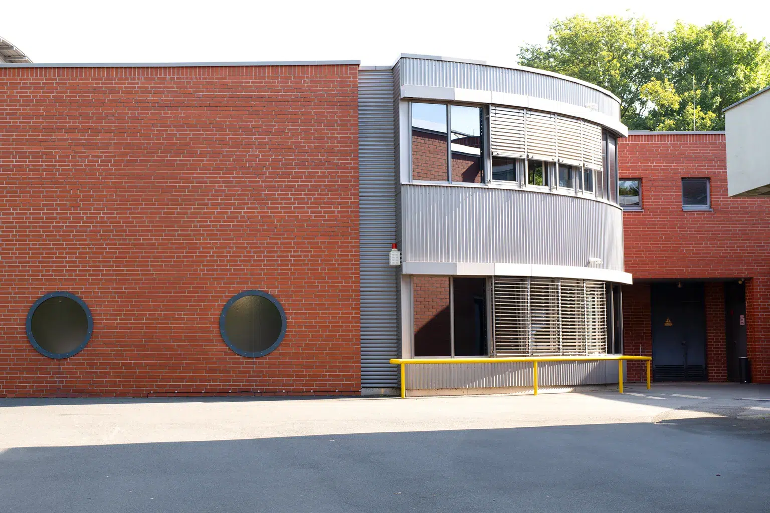 Moderne Gebäudefassade aus rotem Backstein mit runden Fenstern und einem geschwungenen Gebäudeteil aus Wellblech.