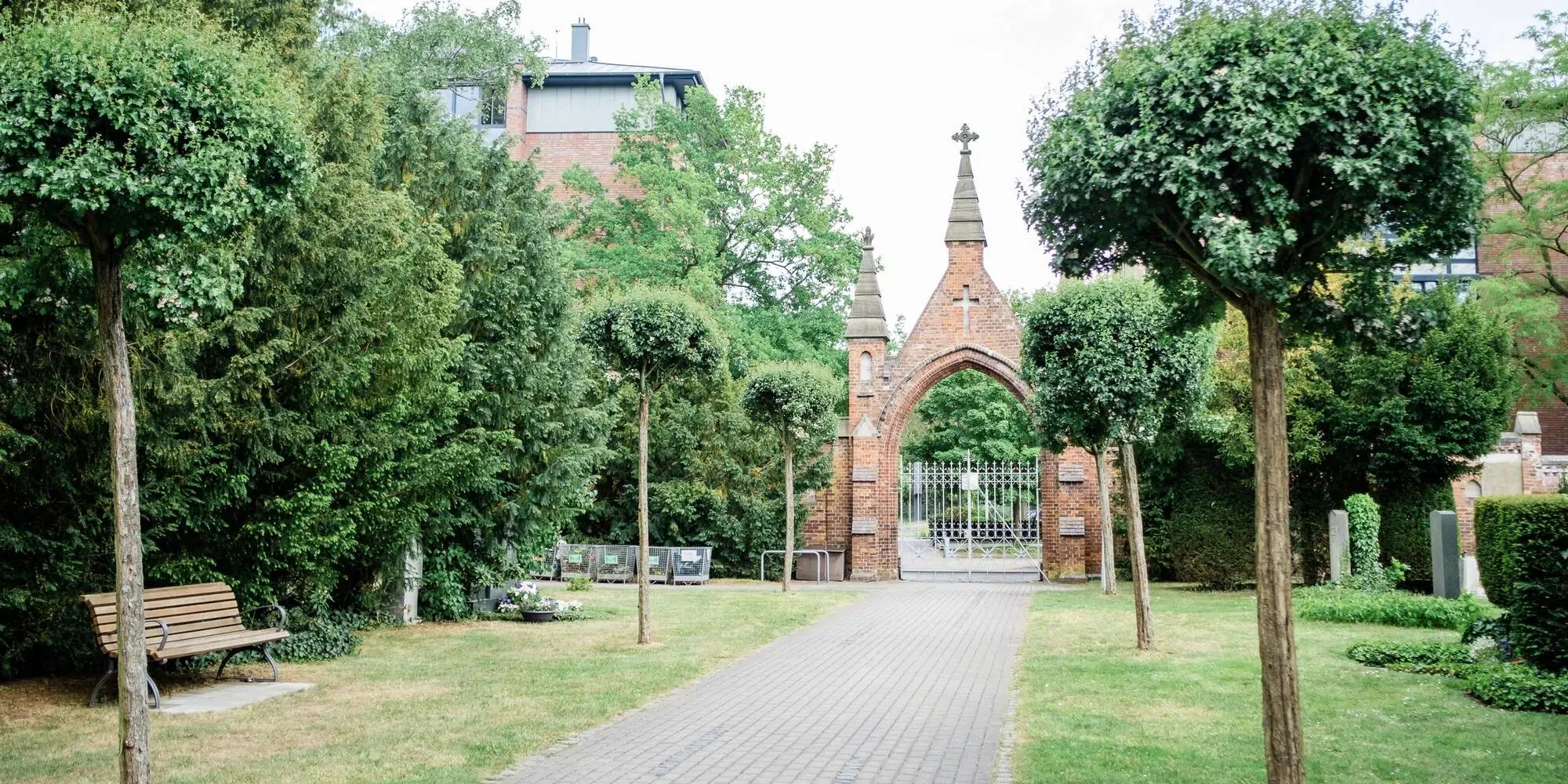 Ein gepflasterter Weg führt durch eine grüne Friedhofsanlage mit Bäumen und einer Bank zu einem historischen Backsteinbogen mit Gittertor.